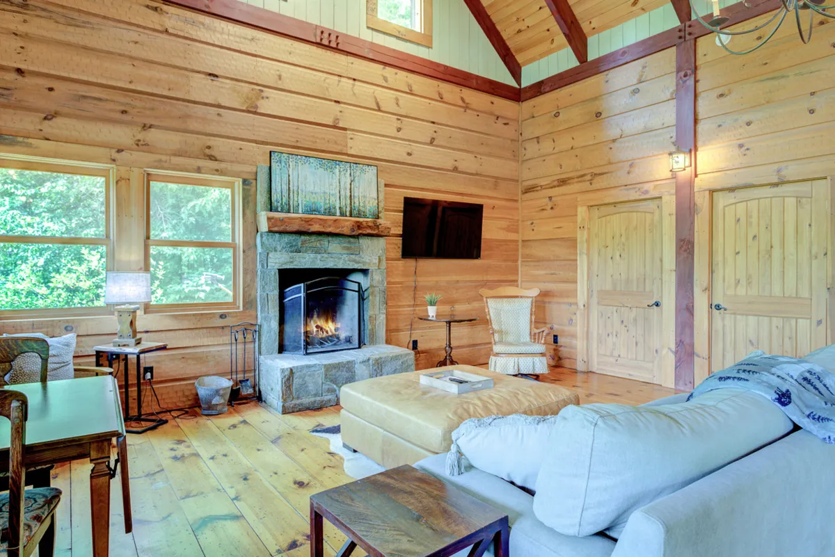 Inside the cottage, a cozy sofa sits by a crackling fireplace against warm wooden walls, with large windows that let in natural light and offer a glimpse of the outdoors. The space feels inviting and comfortable, ideal for socializing or relaxing after a day full of activities.