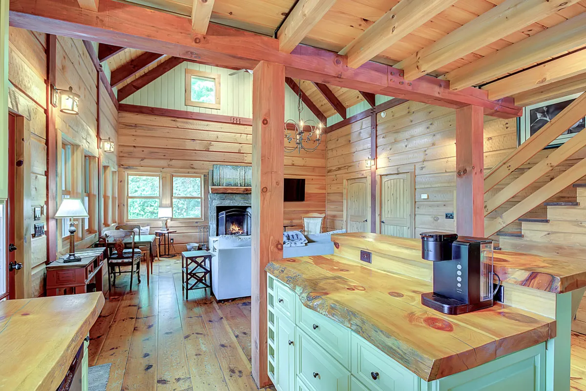 A wide-angle view of the kitchen island, fireplace, and dining area all in one shot. The kitchen island invites gathering, while the adjacent living and dining spaces allow easy interaction. Warm lighting, a visible staircase, and a cozy fireplace complete the inviting atmosphere.