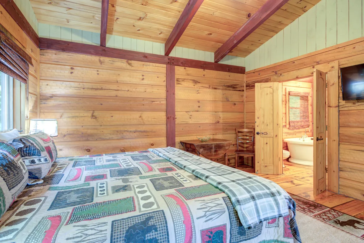 A charming bedroom under rustic wooden ceilings, with a bed dressed in patterned bedding, a small table and chairs for relaxing or playing games, and a doorway leading to a bathroom. The loft-like design and open feel create a cozy but semi-private space.