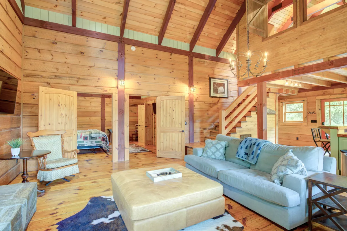 This image captures a spacious living room with a prominent sofa, a striking chandelier, exposed wooden beams, and an adjacent staircase. Through the open layout, the dining area is visible, and large windows bring in abundant natural light. The warm, wood-accented interior and comfortable furnishings evoke a sense of togetherness and relaxation.
