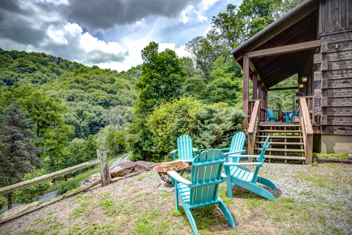 A set of blue Adirondack chairs encircle a fire pit on a spacious lawn, with wooden steps leading up to the house and tall trees in the background. The setup is ideal for gatherings, stargazing, and storytelling around the fire under a mountain sky.