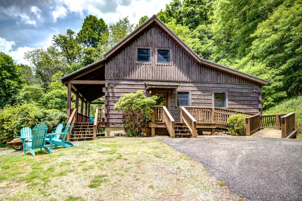 This image features the exterior of the Cozy Mountain Cottage, framed by lush greenery and welcoming Adirondack chairs on the porch. The wooden home radiates a warm, inviting atmosphere, suggesting space for outdoor gatherings and showcasing the natural beauty that surrounds the cottage. The setting sun hints at relaxing evenings and fun family memories.
