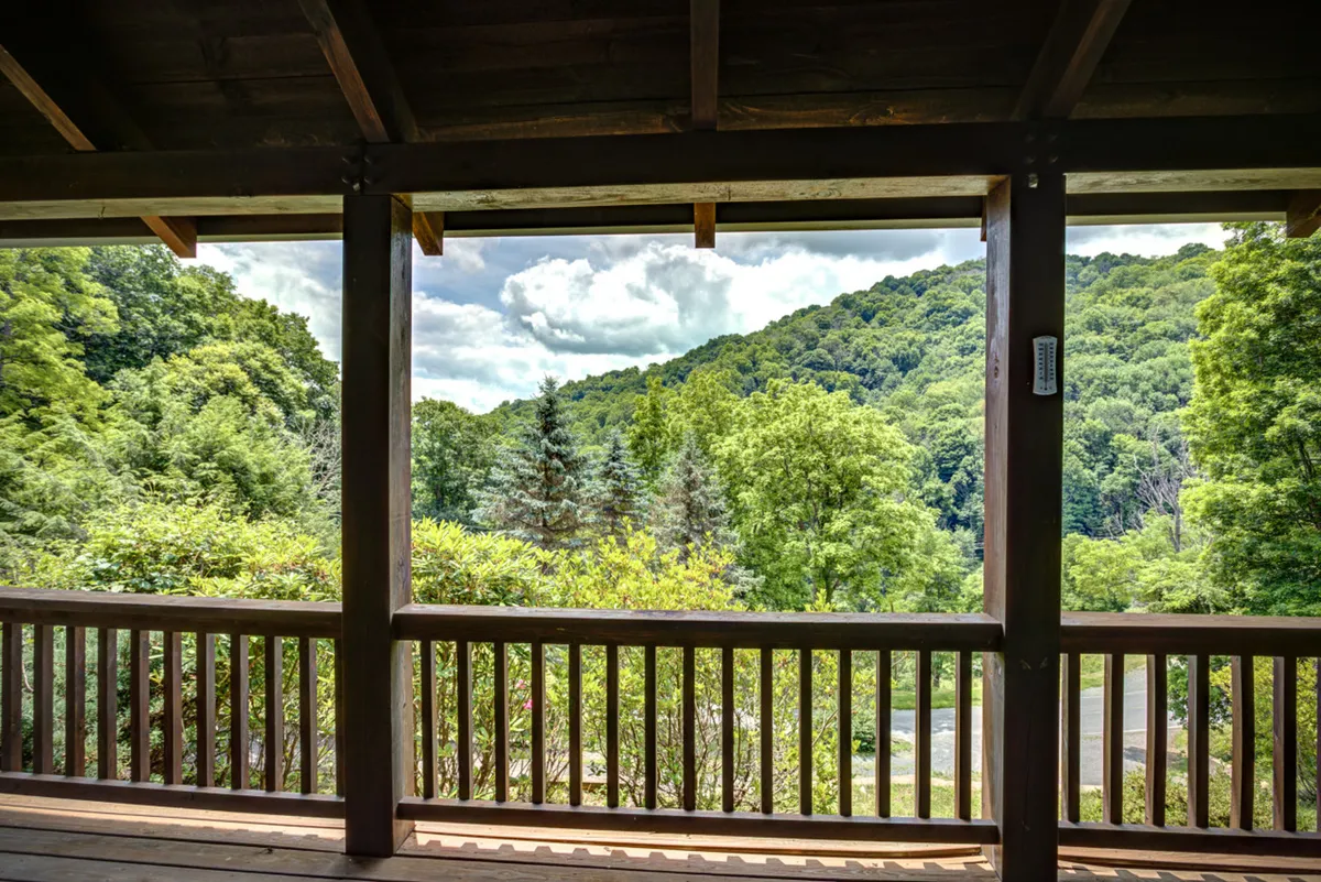 A tranquil view from the porch includes a sturdy wooden railing, lush greenery, and distant blue mountains beneath a cloudy sky. The scene radiates peacefulness and connection to nature, suggesting a perfect spot for quiet moments together.